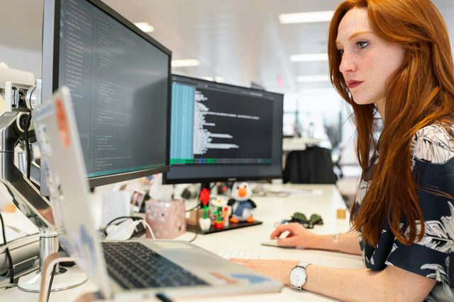 Engenheira de software ruiva concentrada diante de dois monitores com código e um notebook, num escritório claro